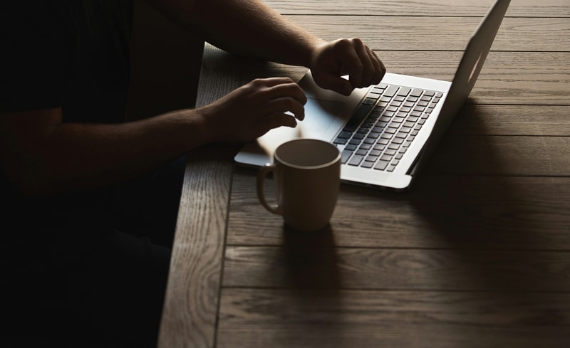 A person sits at a wooden table typing on a silver laptop with a beige mug placed in front, illuminated by soft natural light from the right.