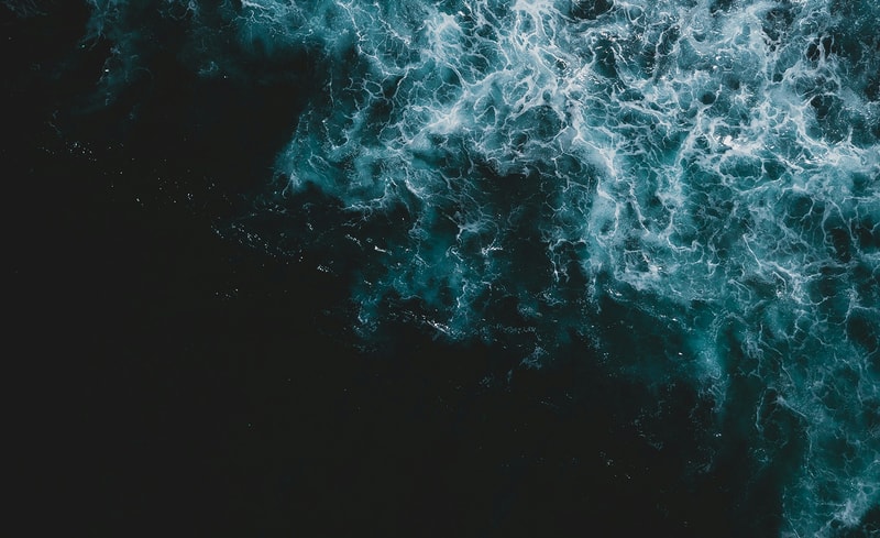 Aerial view of dark ocean water with teal waves and white frothy surf along the right side, contrasting with the nearly black left portion.