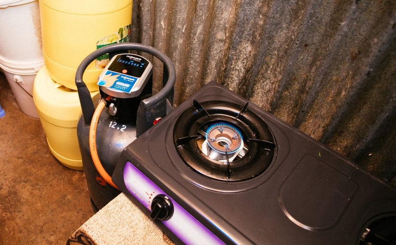 A portable gas stove with a blue flame on a single burner sits next to a gray LPG cylinder and a yellow plastic container in a cluttered, industrial setting.