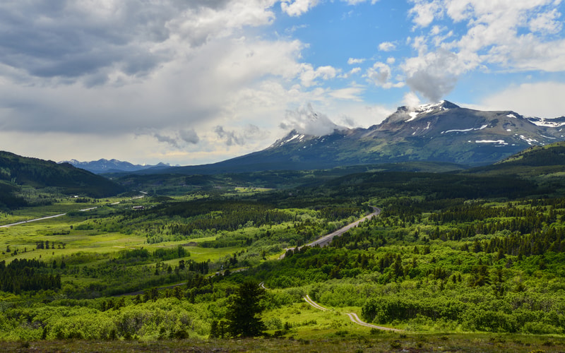 Rugged mountain range with snow patches, lush green valley, winding roads and dense forests under a partly cloudy sky.