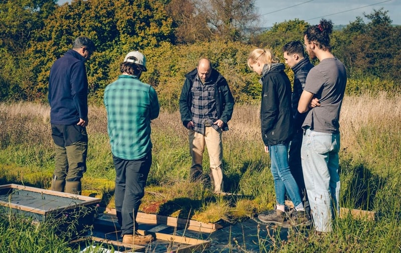 Group of six people standing outdoors in a grassy field, gathered around a grid with plants; one man in center appears to explain or inspect, others watch attentively.