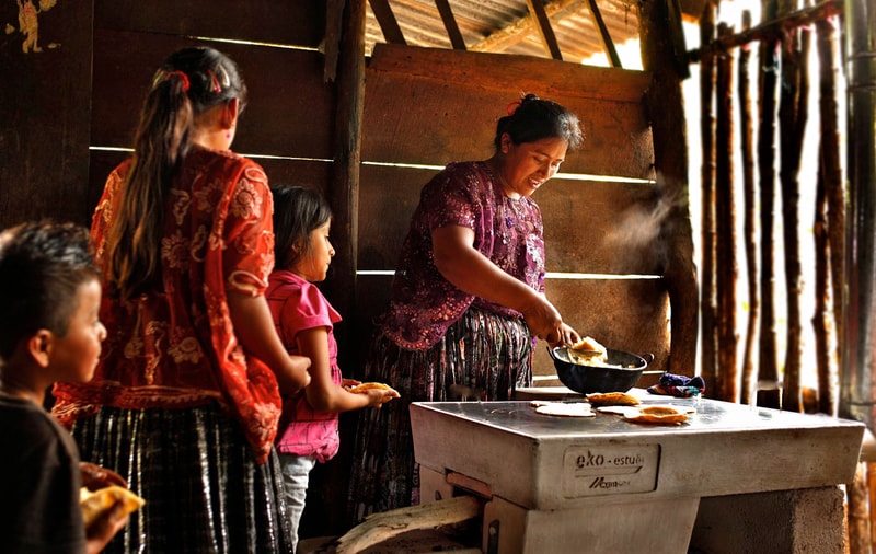 A woman stirs food in a frying pan while several children stand in line, watching inside a rustic wooden kitchen with warm, natural light.