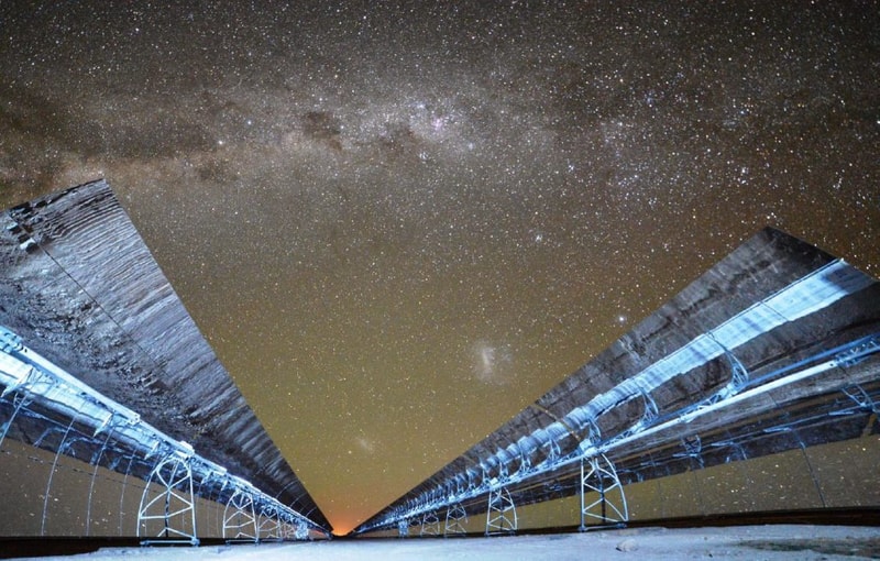 Long-exposure view of a starry night sky framed by two parallel, raised solar panel arrays or rails converging toward a distant orange horizon.