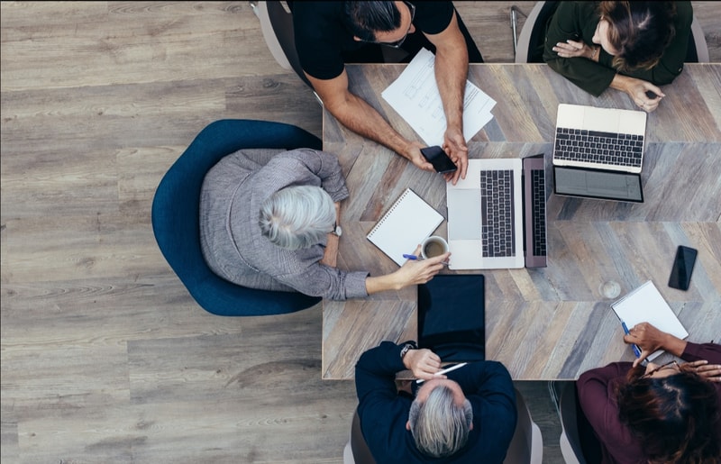 A top-down view of a diverse group of people around a wooden conference table with laptops, tablets, notebooks, mugs, and devices, collaborating and taking notes.