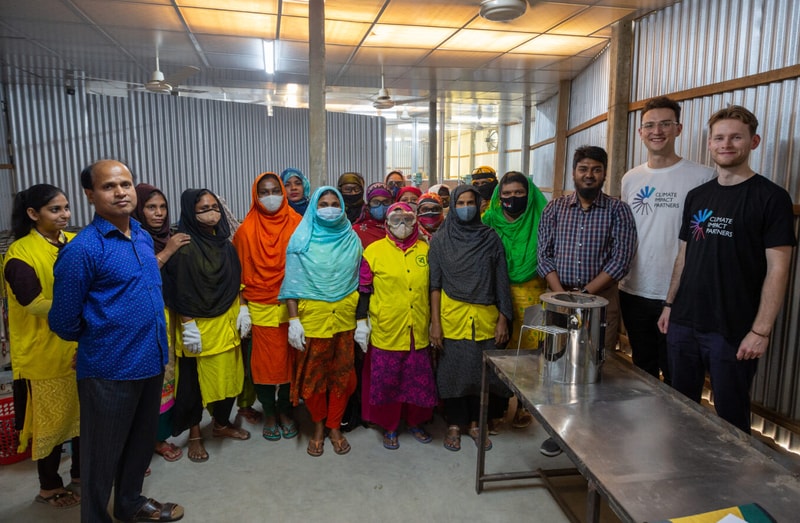 A diverse group of people, many wearing colorful traditional clothing and headscarves, stand together in a metallic workshop with a stainless table and equipment.