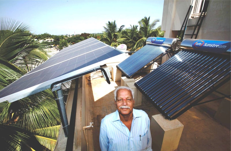 Sunlit rooftop with multiple solar water heaters and solar panels; a smiling older man in a light blue striped shirt stands in foreground, palm trees in background.