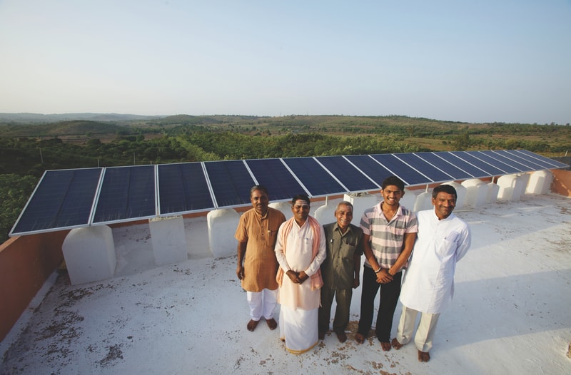 Group of five men standing on a white rooftop beside a row of solar panels, with a green landscape in the background under a clear sky.