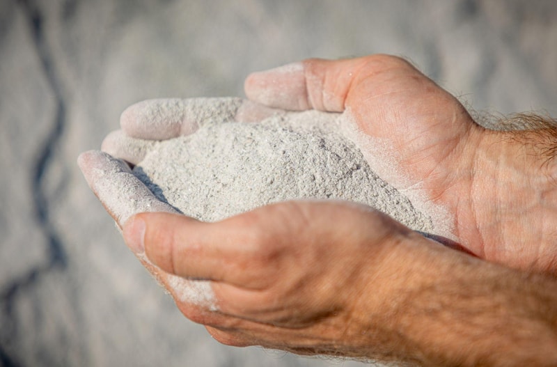 A pair of weathered hands cupping a handful of fine white powdery substance, with powder dust on fingers and palm, light gray background.