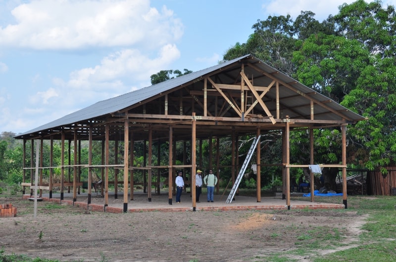 A wooden open-frame pavilion under construction with a metal roof, several workers inside, a ladder resting against a beam, and trees in the background.