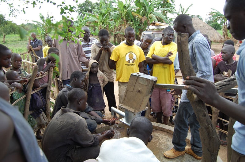Group of people gathered around a hand-operated water pump in a rural area, with children and adults watching as water flows into a basin.