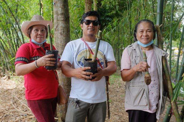 Three people stand outdoors among green trees, each holding a small potted plant with a bamboo stake; one wears a hat, another sunglasses, all with masks hanging.