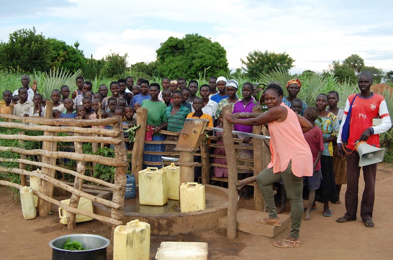 Crowd of villagers gathered around a wooden water pump and jerry cans, listening to a woman in a peach top as she operates the pump in an open rural setting.
