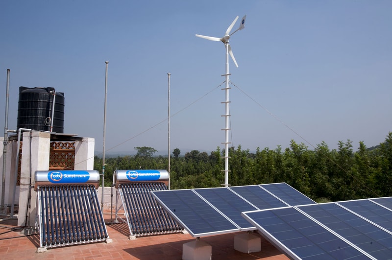 Rooftop solar thermal tubes and solar panels on a terrace with a wind turbine, water tank, and wires against a clear blue sky and green distant trees.