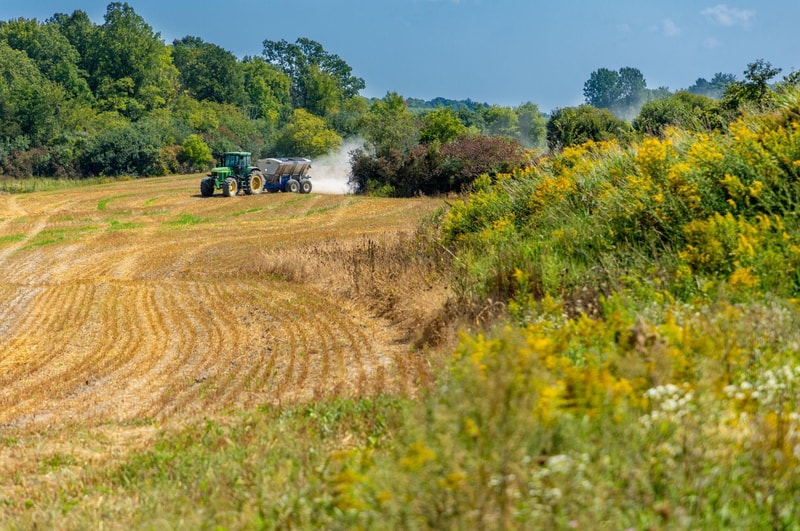 Tractor pulling a trailer across a harvested field, kicking up dust with a row of yellow wildflowers and green trees in the background under a blue sky.