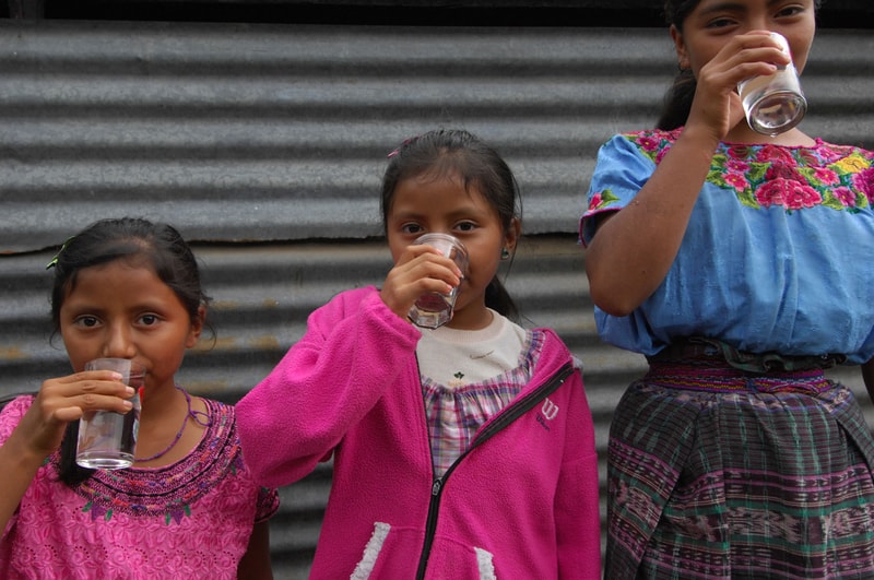 Three young girls stand in front of a corrugated metal wall, each drinking from a clear glass cup, wearing colorful traditional and casual clothing.