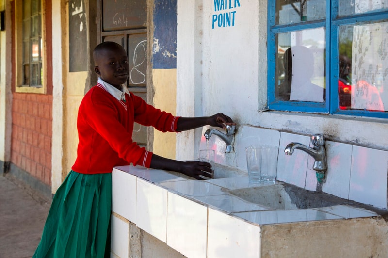 A young person in a red sweater and green skirt taps a water faucet at a tiled outdoor sink, filling a glass beside a blue-framed window on a sunlit street.