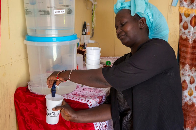 A woman in a turquoise headwrap fills a white paper cup with water from a large dispenser, standing at a red-draped table indoors.