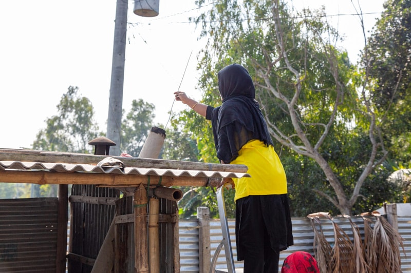 A person wearing a yellow top and black hijab stands on a ladder, reaching up to adjust a long cable or wire near a makeshift rooftop with metal and wood, in a sunny outdoor setting.