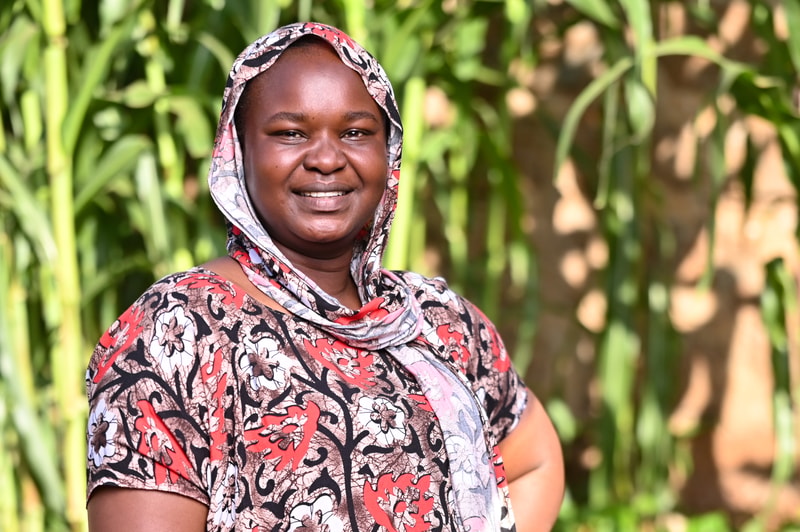 Smiling person wearing a patterned headscarf and matching blouse outdoors, with green plants and blurred brown background behind.