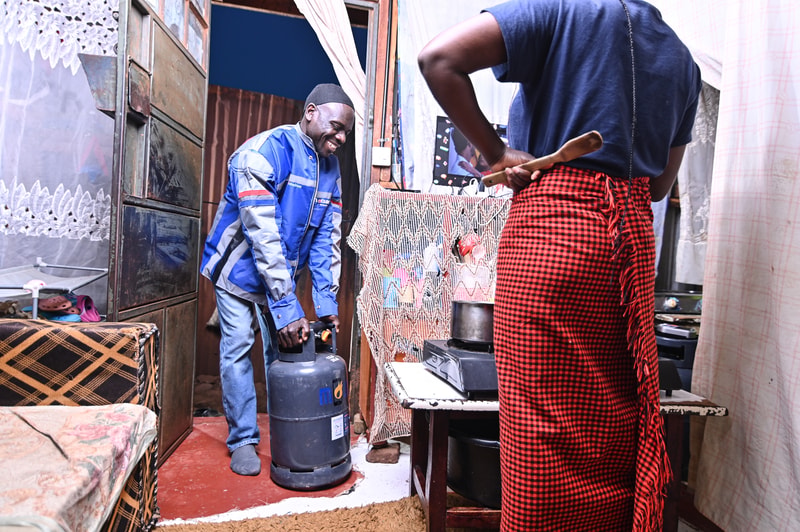 Two men in a cozy, cluttered room: one wearing a blue jacket and cap handles a gas cylinder; the other, with a red plaid wrap skirt, stirs a pot at a table.