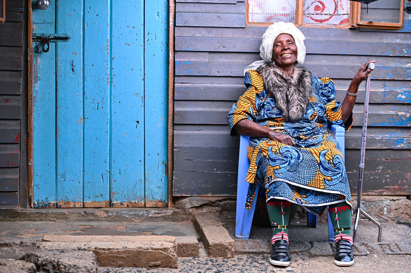 An elderly person wearing a vibrant patterned dress, fur collar, and a white headwrap sits on a blue chair outside a weathered wooden building, smiling, with a cane.