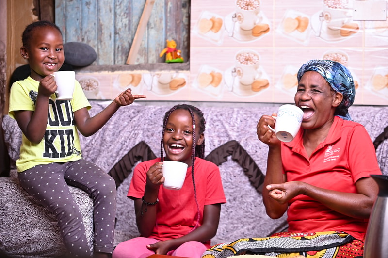 Three smiling people enjoy mugs of tea together: a young girl in a lime shirt on the left, another child in red center, and an older woman in red on the right, indoors.