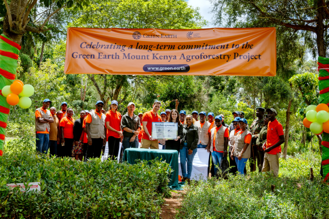A group of diverse people in bright shirts pose under an orange banner that reads “Celebrating a long-term commitment to the Green Earth Mount Kenya Agroforestry Project,” outdoors.