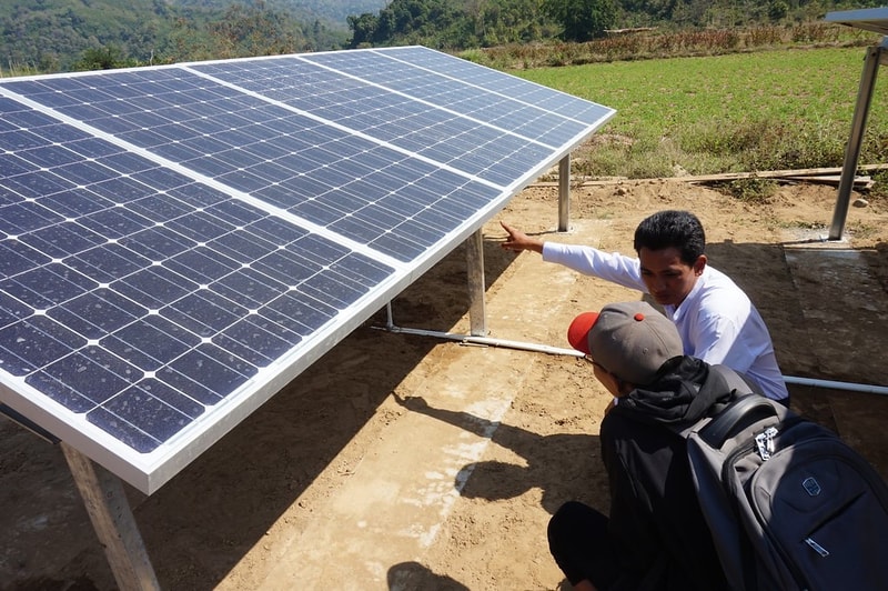 Two people inspect a large solar panel array mounted on metal supports in an outdoor rural area, with one person pointing and the other wearing a backpack.
