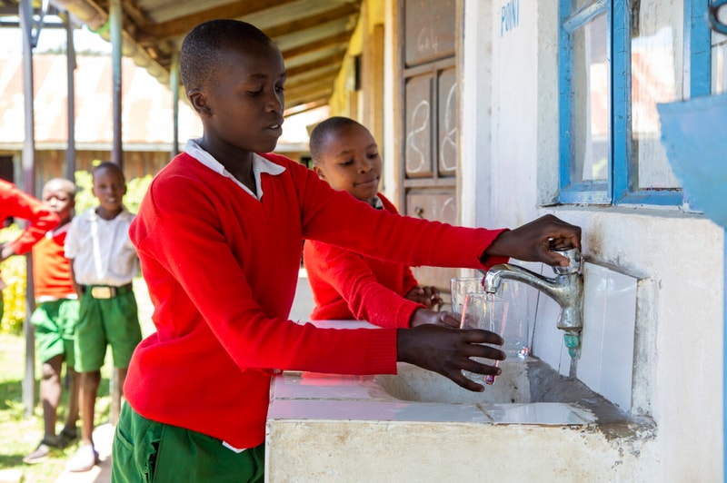 Several young students in red uniforms wash glasses at a shared outdoor sink, filling cups from a running faucet under a sunlit covered walkway.