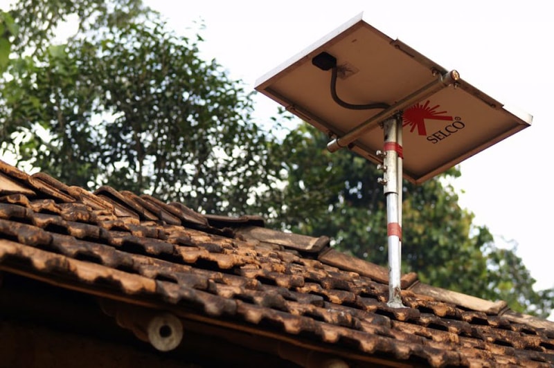 Solar panel mounted on a metal pole secured to a tiled roof, with a visible cable and branding; green trees and bright sky in the background.