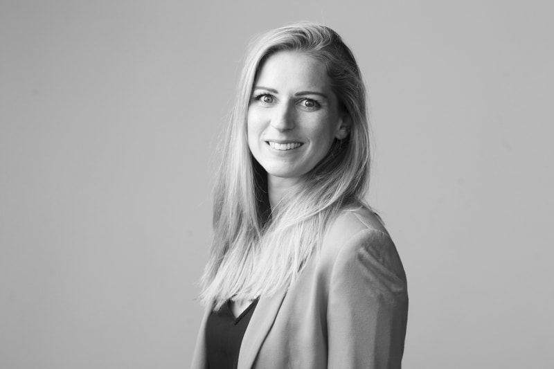 Professional head-and-shoulders portrait of a smiling woman with long blond hair, wearing a blazer over a dark top, facing slightly to the right against a plain background.