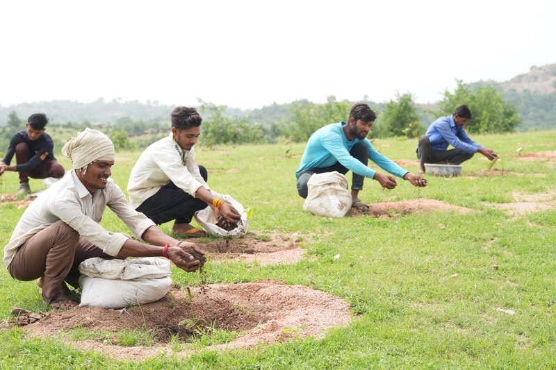 farm workers kneeling in a field, scooping soil and planting seeds into small mounds, wearing casual clothes and head coverings, with bags nearby and green hills in distance