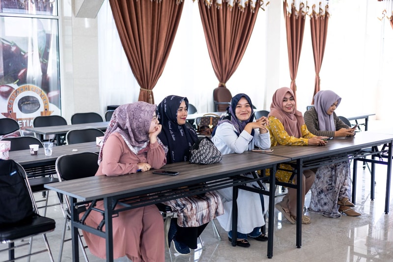 A group of five women wearing hijabs sit at a long classroom table in a bright room with brown curtains, smiling and chatting, some with phones and bags nearby.