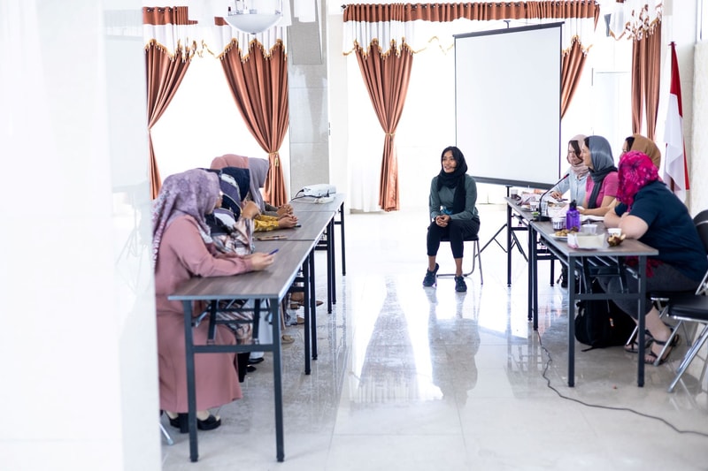 A group of women wearing headscarves sit around long tables in a bright room with brown curtains, a projector screen, and Indonesian flag in the background.