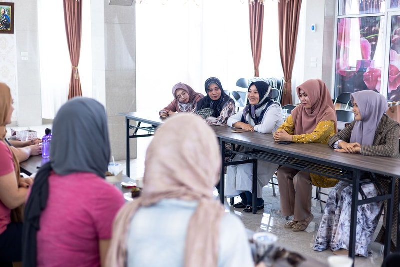 Group of women wearing hijabs seated around a long table in a bright room, chatting and smiling during a informal meeting or gathering.