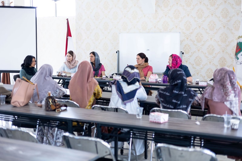 Group of women in headscarves seated at a long meeting table in a classroom or conference room, engaging in a panel discussion with a whiteboard and projector screen in the background.