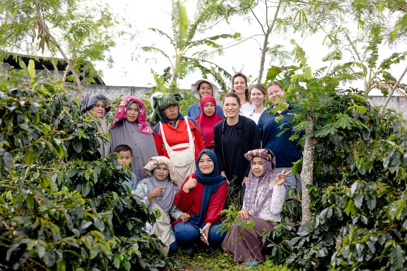 A diverse group of people, including women in headscarves and casual clothing, stand together in a lush garden with trees and plants, posing for a photo.