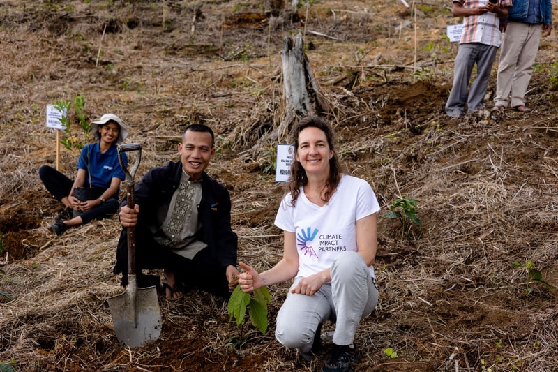 Group of volunteers planting saplings on a hillside; a smiling man holds a shovel beside a woman in a white T-shirt with a Climate Impact Partners logo, kneeling with a plant.