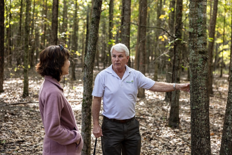 Two adults conversing in a sunlit forest; an elderly man in a white polo shirt leans against a tree while a woman in a pink jacket listens nearby.