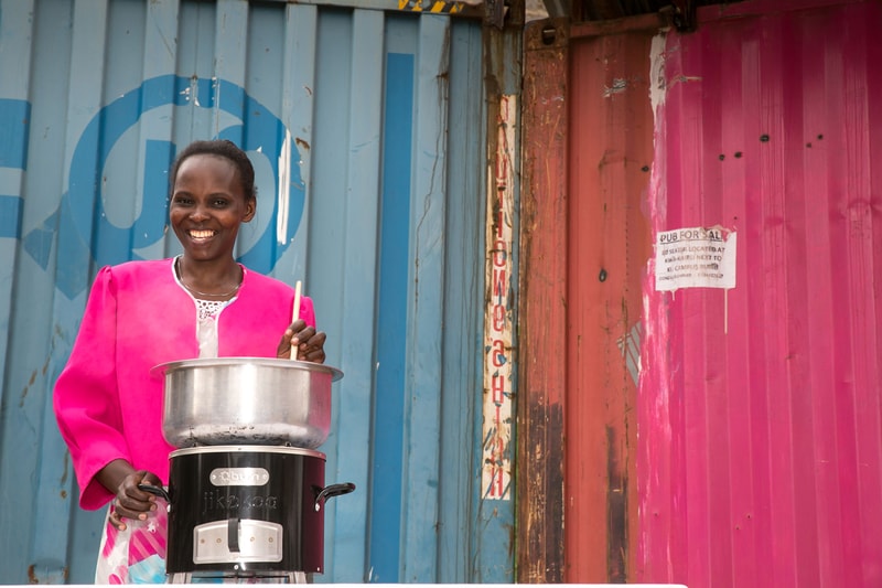Smiling woman in a bright pink jacket stands beside a metal pot atop a black appliance, cooking with a wooden spoon against a colorful backdrop of blue and pink corrugated metal walls.