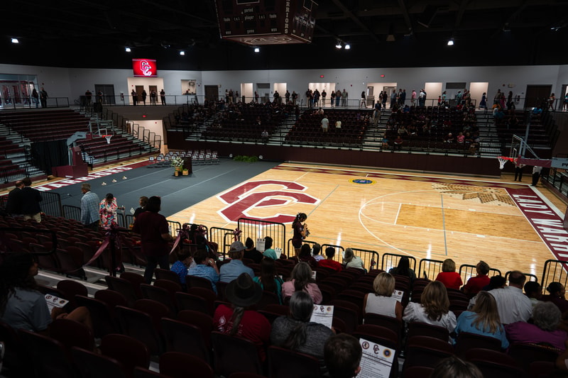 A gymnasium with a hardwood basketball court, large central logo, bleachers filled with spectators, and people walking along upper concourse.