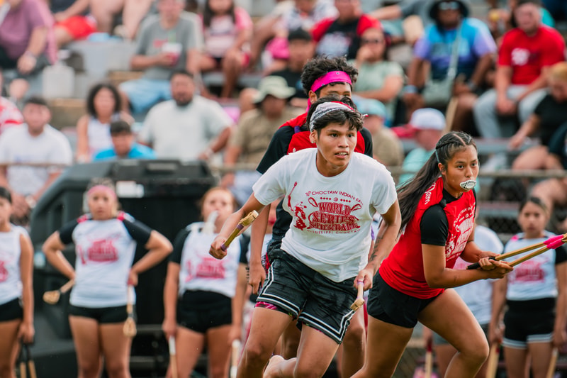 Young participants compete in a stickball race at a crowded outdoor event, sprinting forward with sticks while spectators watch from bleachers in the background.