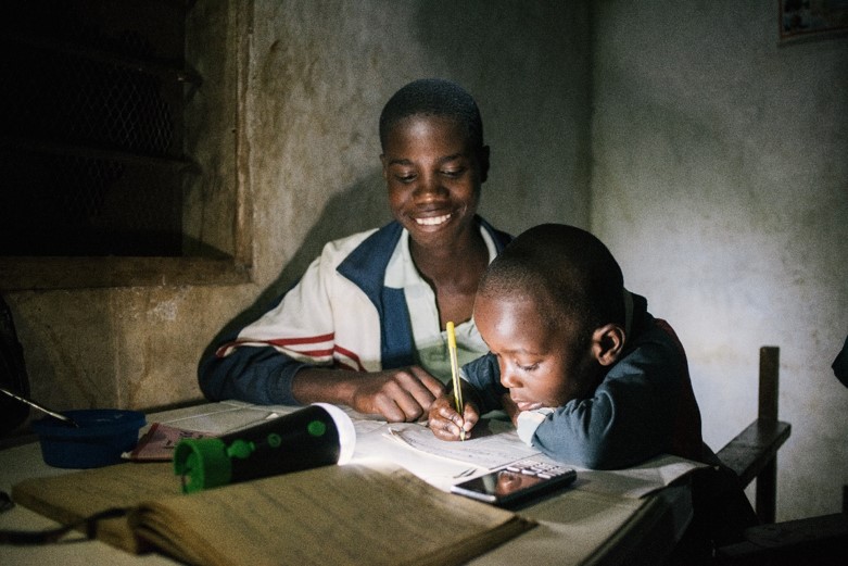 Two young people study at a wooden table, one smiling while guiding the other who writes with a pencil; books, a phone, and a flashlight sit nearby in a dim room.