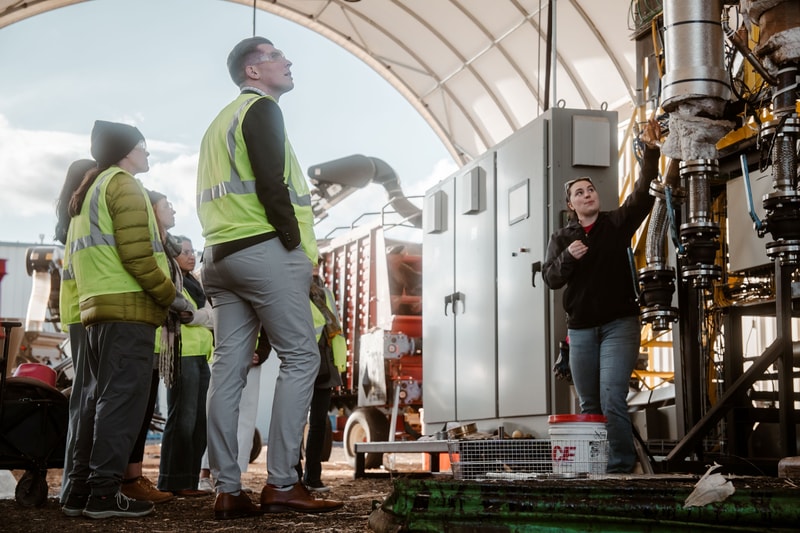 Group of workers in high-visibility jackets listen to a technician explaining large industrial equipment under a curved shelter.