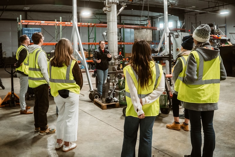 A group of people in high-visibility vests listening to a woman in a black jacket giving a briefing inside an industrial workshop with machinery, shelves, and concrete floor.