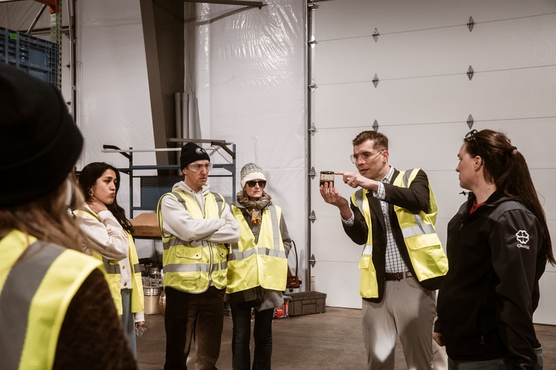 Group of people in high-visibility vests listening to a man in a suit who demonstrates something on a small device inside a warehouse or workshop.