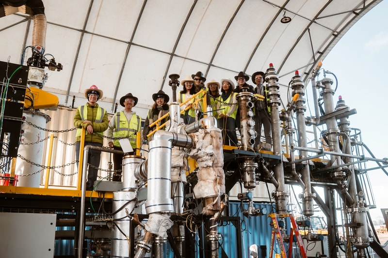 Group of workers in safety vests and hats stand on an industrial platform among large metallic pipes and equipment under a curved canopy.