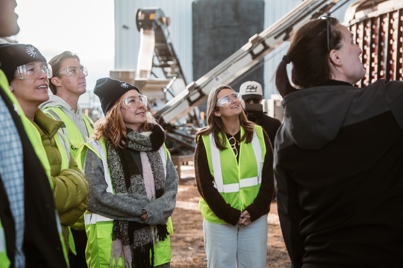 Group of people in safety vests and goggles on a construction site, listening to a guide near industrial equipment and a conveyor system.