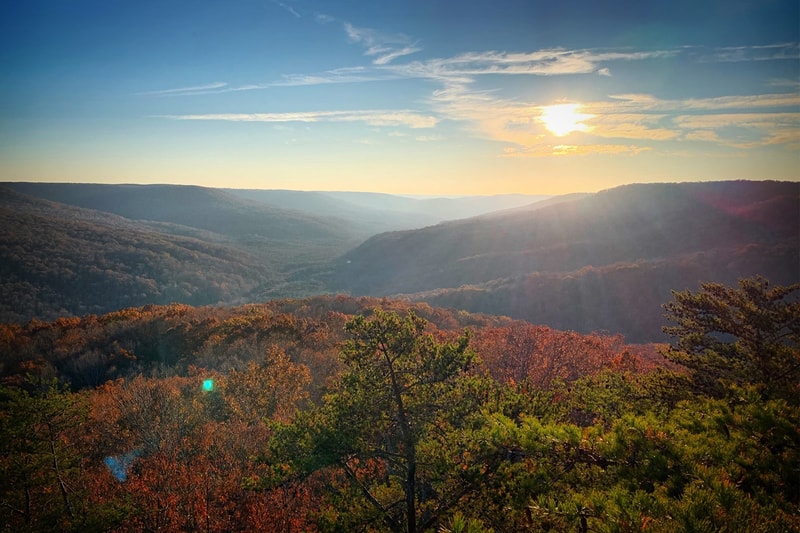 Sunlit valley and rolling hills during autumn, with a bright sun low in the sky, layered blue ridges, and colorful orange-green forest in the foreground.