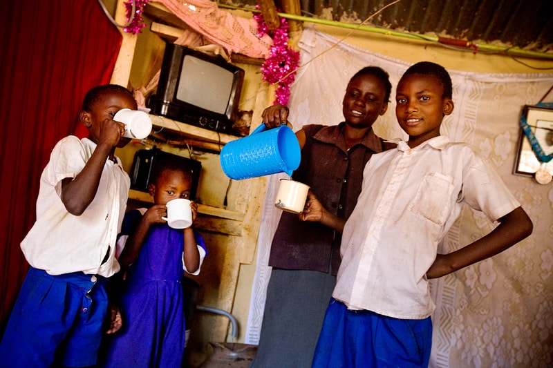 Four children stand indoors, one pours from a blue cup into a beige mug while others drink from white mugs; a woman watches in the background, festive decorations around.